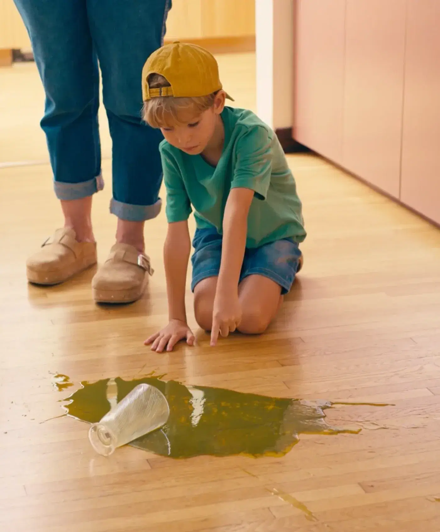 A boy kneeling on a wooden floor points at a spilled drink beside a tipped-over glass