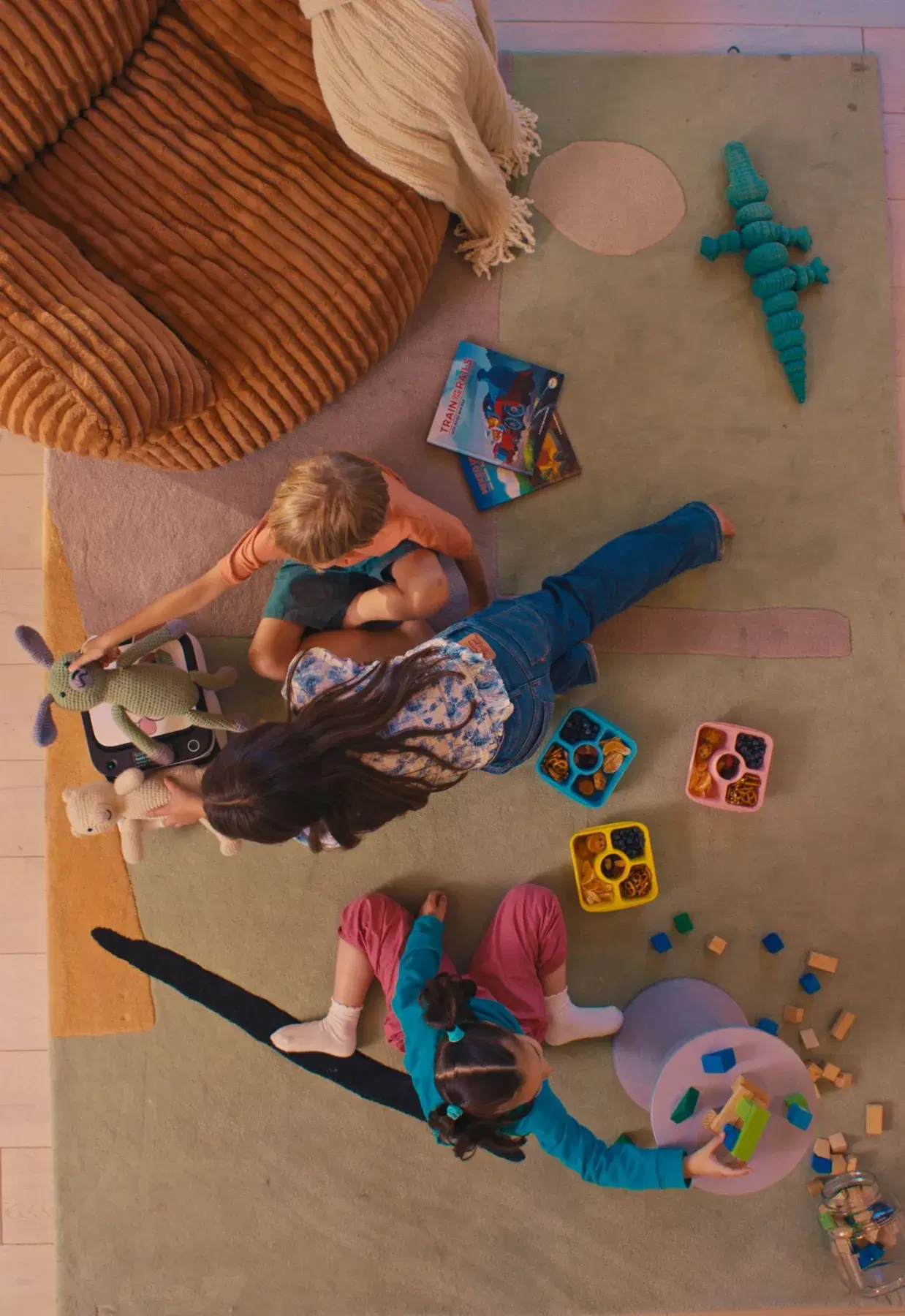 Three children playing on a rug strewn with blocks, books, snack trays and plush toys, seen from above