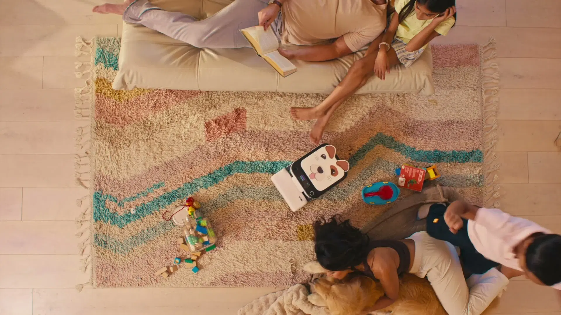 A family relaxes on a rug from above, reading and playing with the dog, while Matic cleans among them