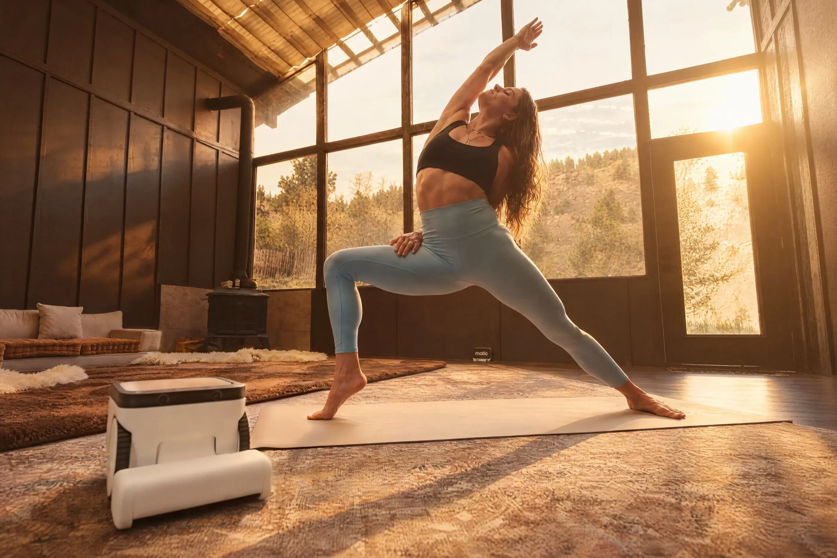 A woman reaches into a side stretch by tall sunlit windows while Matic cleans the rug beside her mat